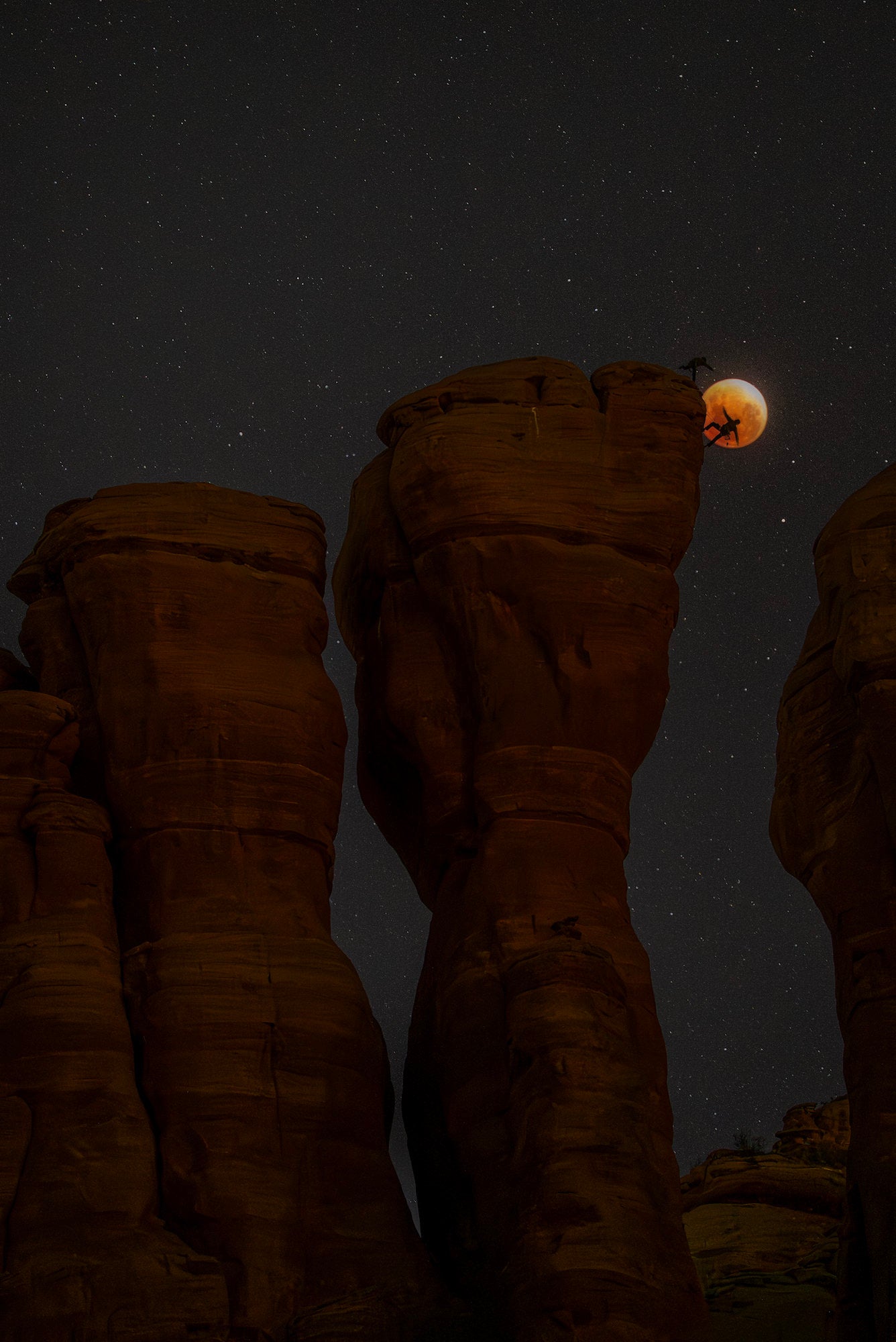 Lunar eclipse framed beside a sandstone tower as two climbers silhouette against the red moon and star field.