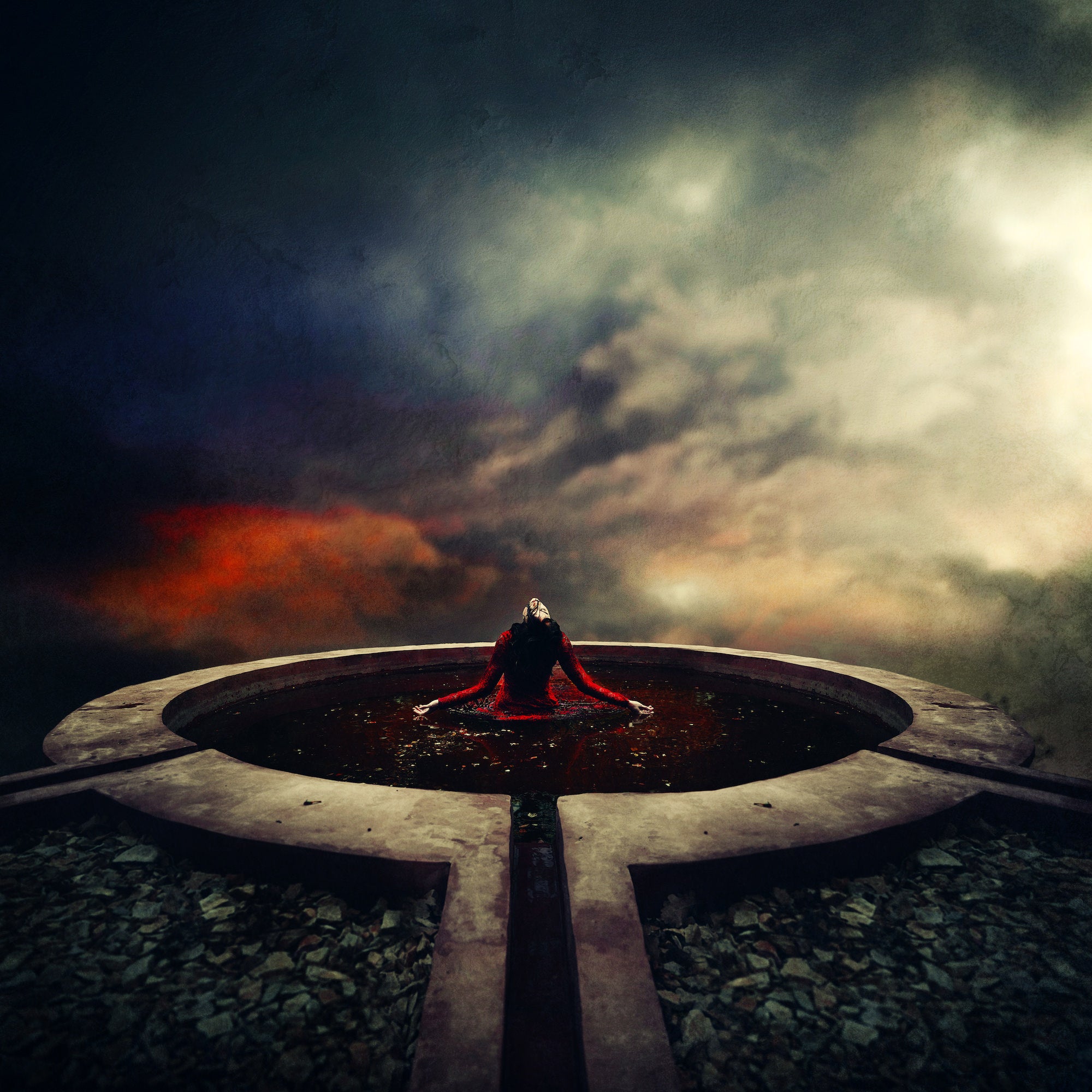 A wide-angle fine art image showing a masked figure in a red lace dress kneeling with arms outstretched in a circular stone fountain, surrounded by dark water and fallen leaves, beneath a moody, atmospheric sky.