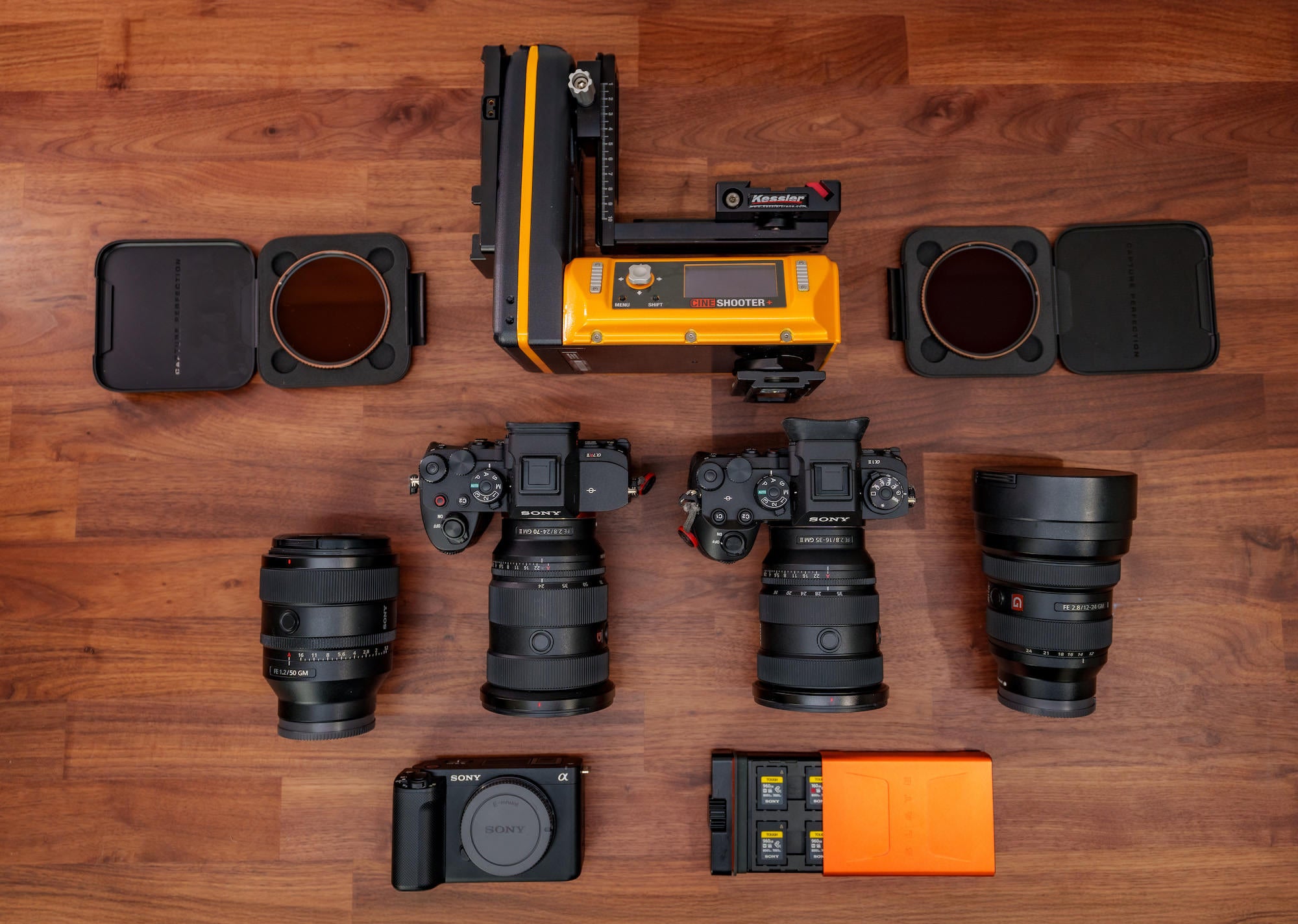 Flat lay of Sony cameras, G Master lenses, motion‑control head, filters, and media cases arranged on a wooden surface.